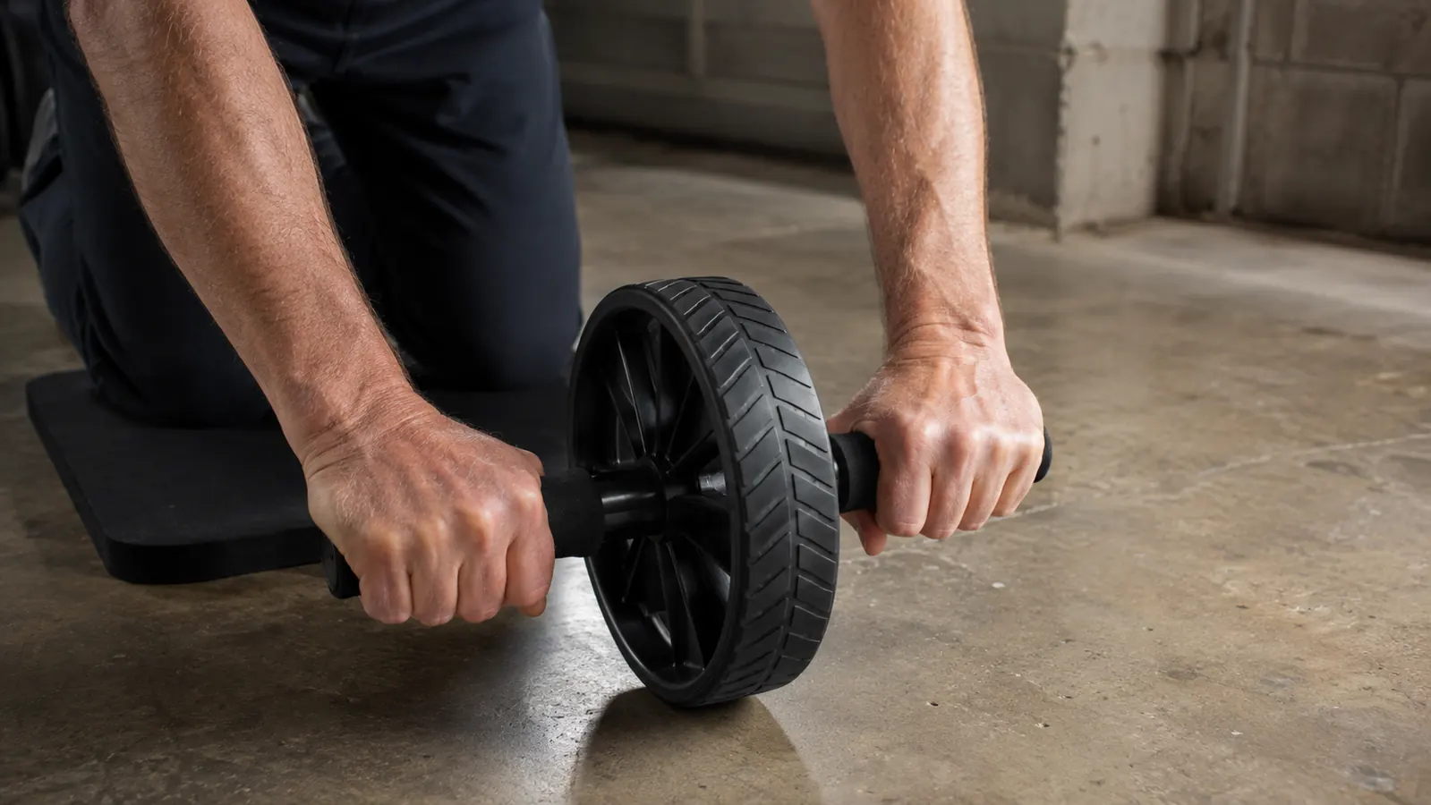 A hand holding the handles of an ab wheel on a garage floor, knuckles down, wheel planted, a knee mat beneath