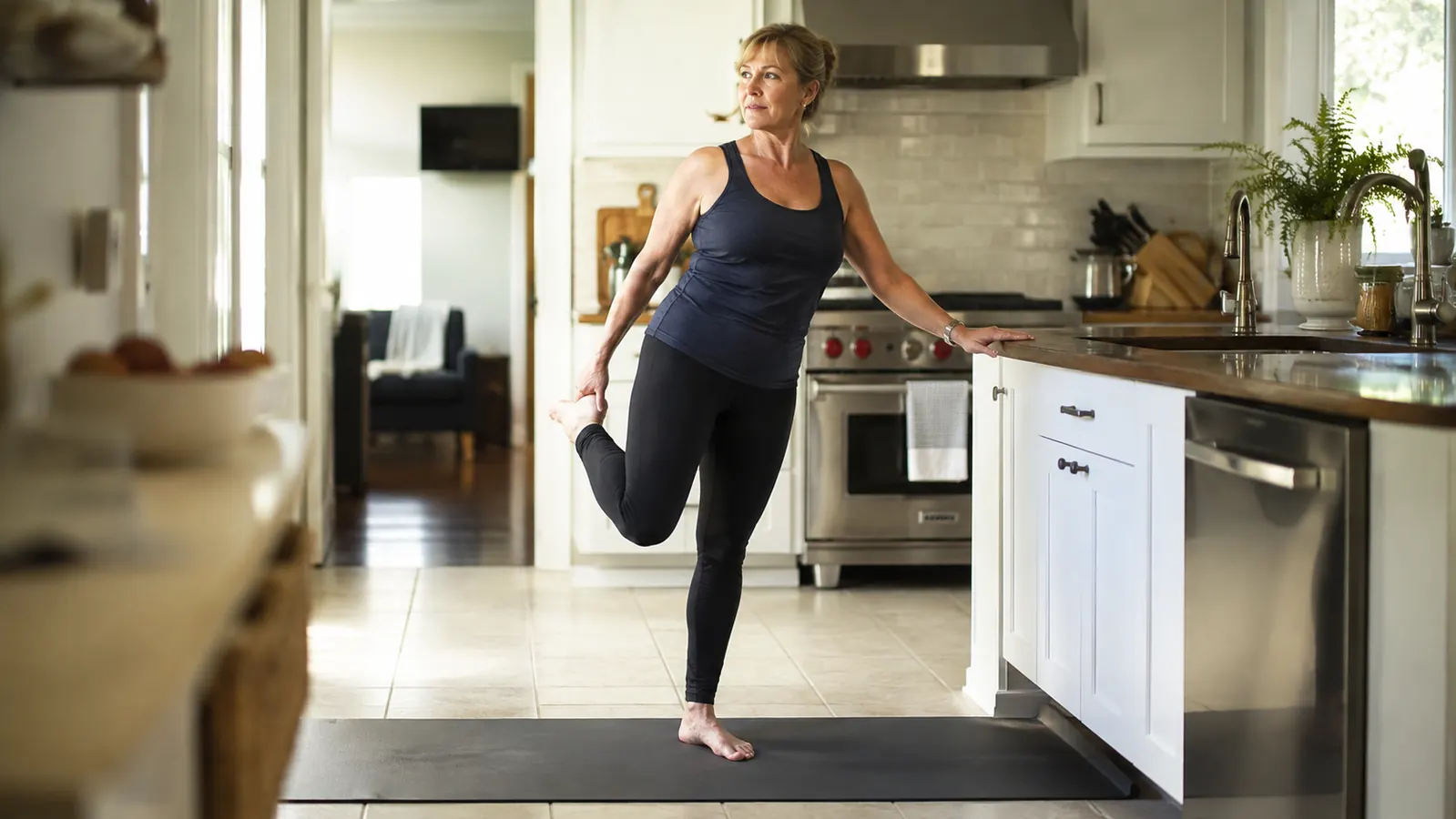 A woman in bare feet standing balanced on one leg on a thick exercise mat in a bright kitchen, a countertop within reach, tiles visible at the edges of the mat