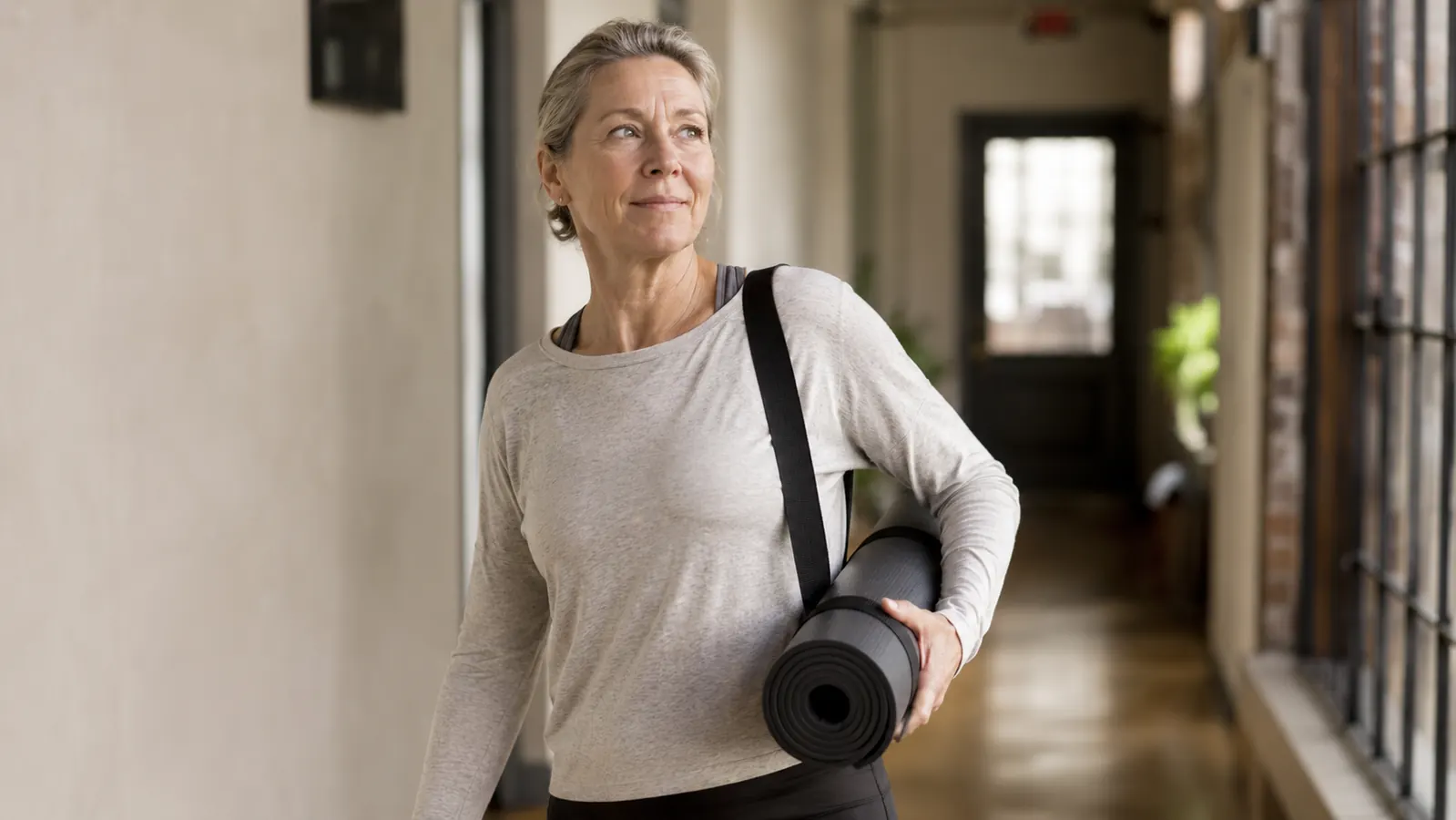 A thick yoga mat rolled up and slung by its carrying strap over a shoulder, held by a hand, in a bright hallway near a door