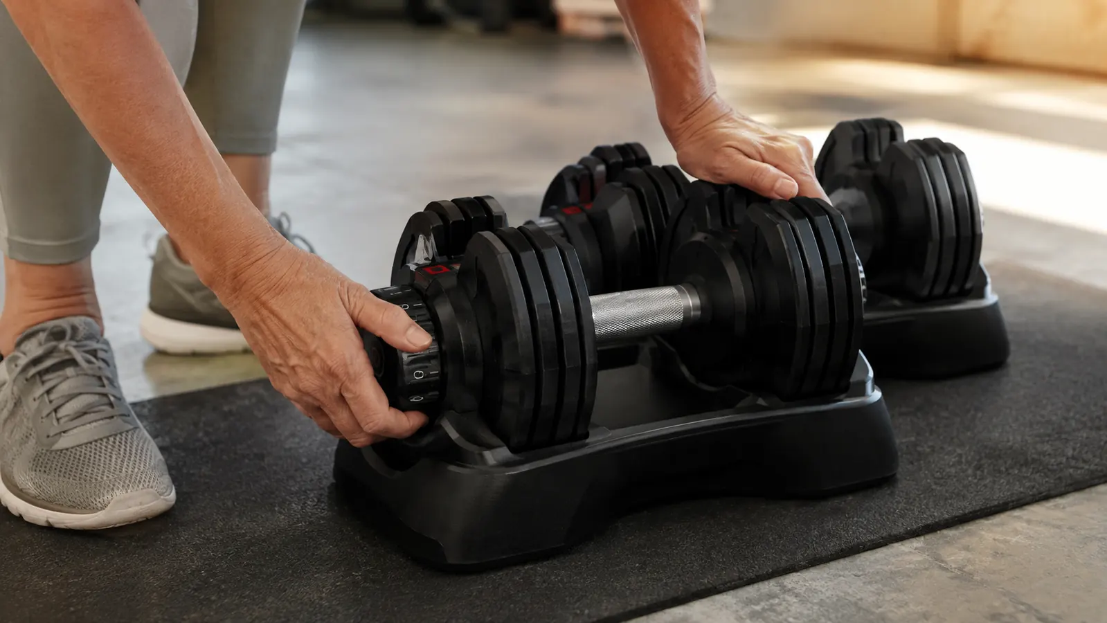 Hands turning the weight dial on a pair of adjustable dumbbells on a garage floor